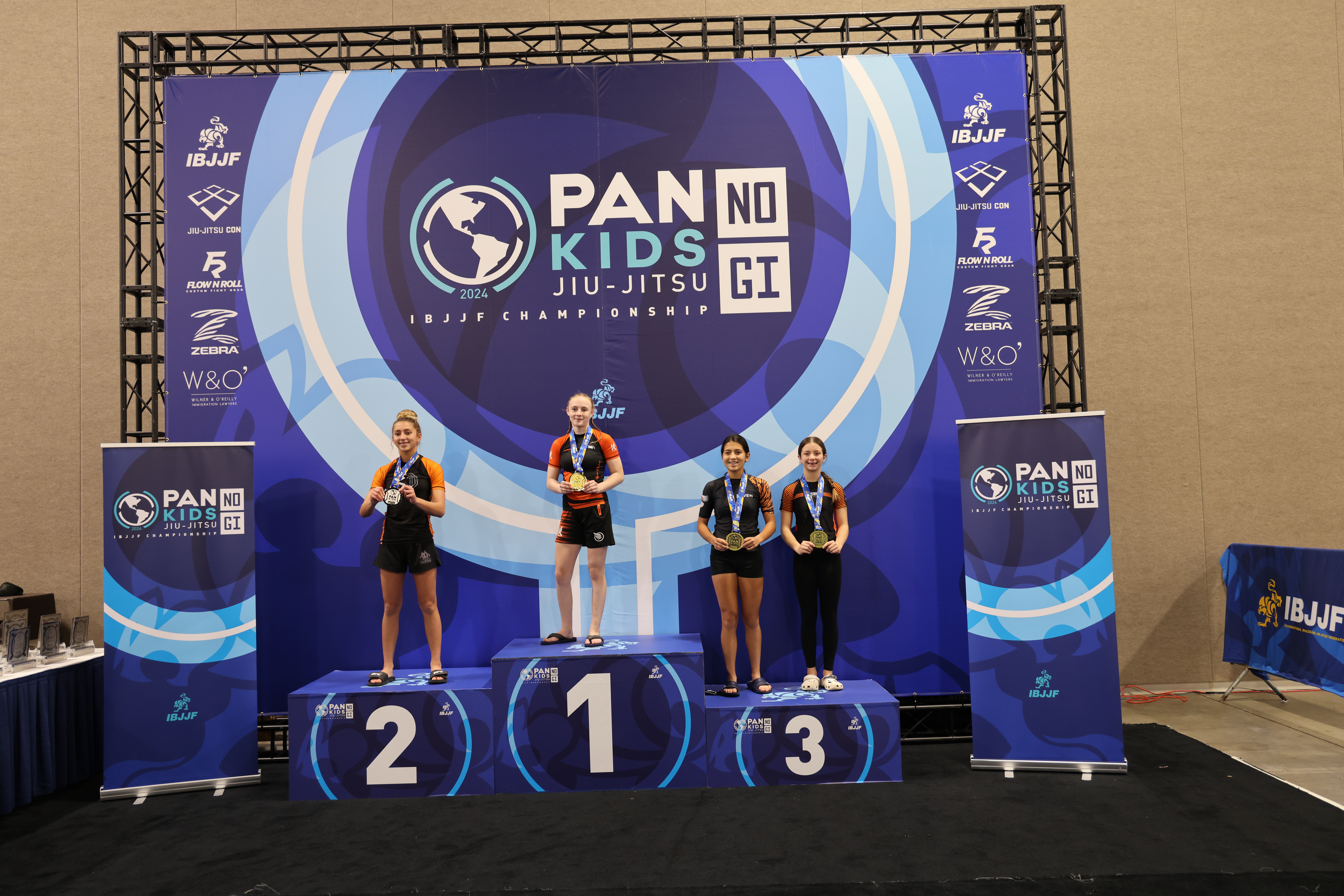 Podium photograph from the 2024 Pan Kids IBJJF No-Gi Championship. The top step of the podium is occupied by the division champion. Event branding is visible in the backdrop: PAN KIDS NO-GI IBJJF CHAMPIONSHIP.